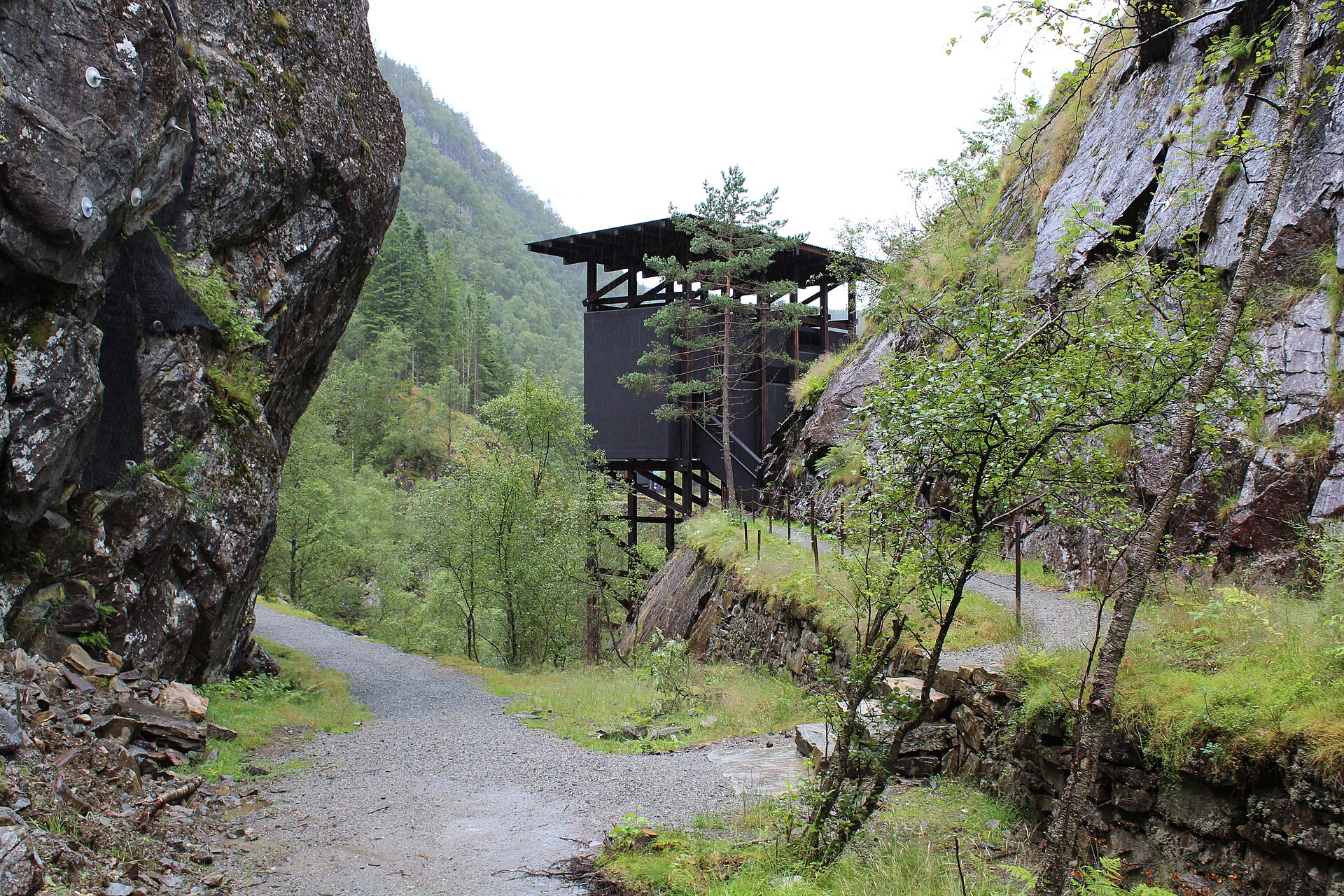 Et mørkt trebygg på påler i et trangt, grønt fjelljuv, med en grusvei i forgrunnen.