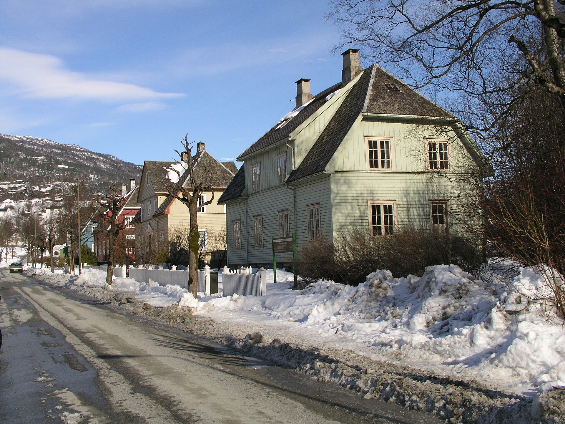 En rekke eldre trehus langs en vintervei, med snø i veikanten og fjell bak.
