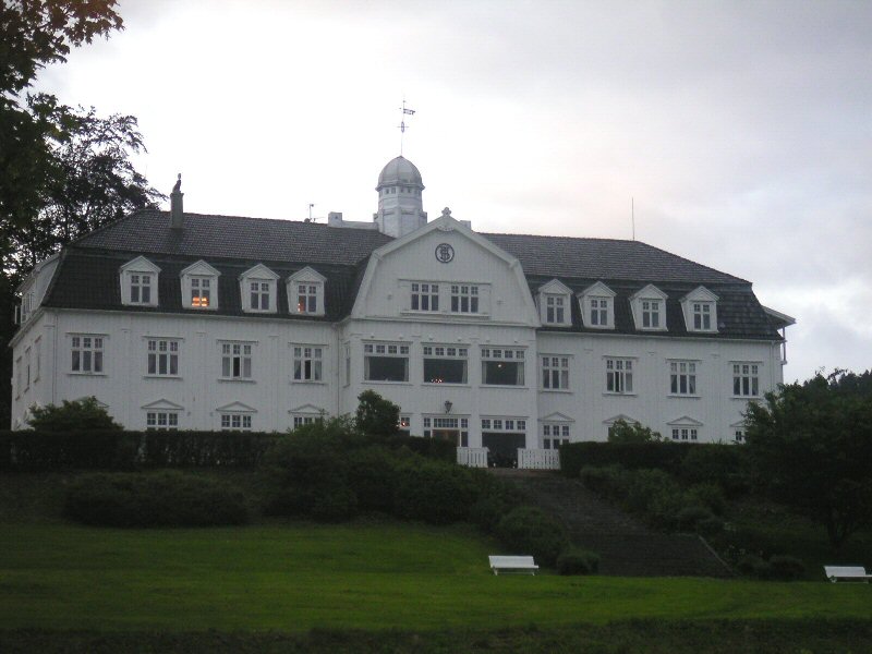 Stort hvitt trehotell med valmtak og et lite tårn, sett over en gressbakke.