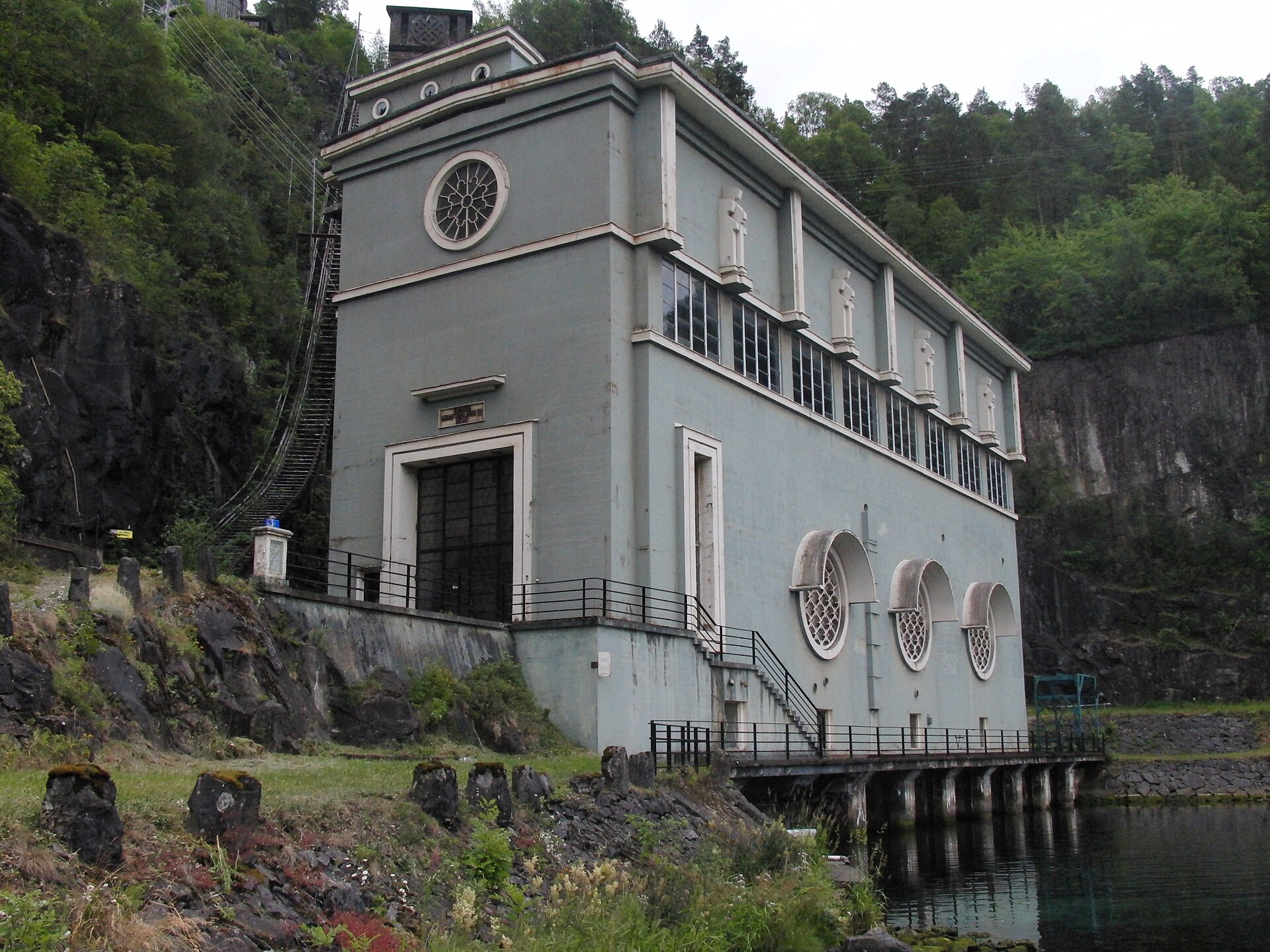 Grå art deco-kraftstasjon med runde vinduer, bygget inn mot en bratt fjellvegg ved vannet.