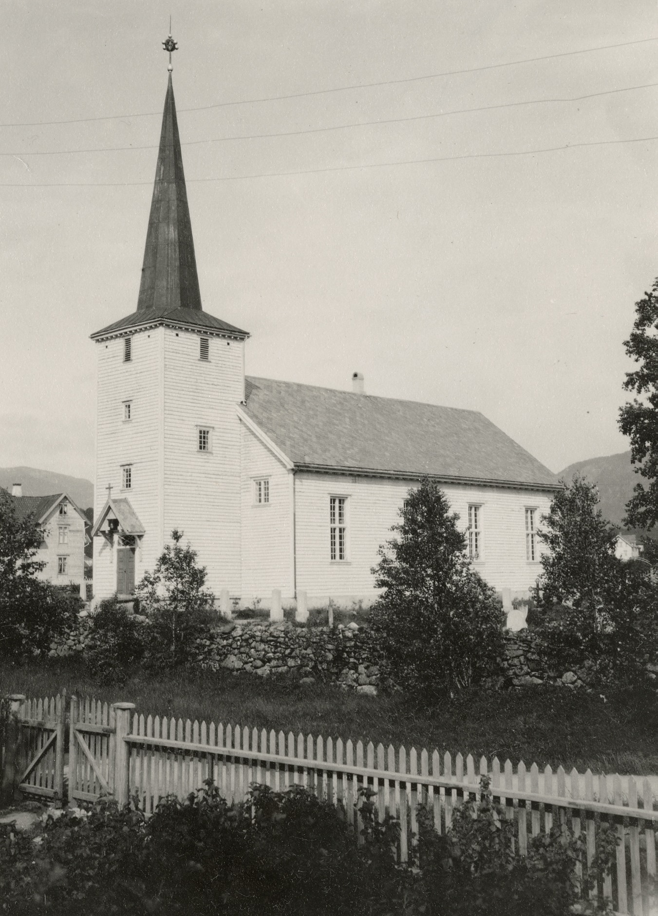 Hvit trekirke med spir, fotografert i snø i 1950, med fjell bak.