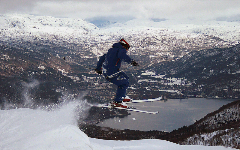 En skiløper i hopp over en snøkant, med Saudafjorden og snødekte fjell bak.